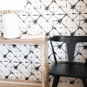 May include: A modern interior scene featuring a white lamp, a decorative knot, and a black chair. The background showcases a white wall with a black geometric pattern. The furniture sits on a light wood table and floor.