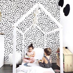 K&ouml;nnte beinhalten: Ein wei&szlig;er, hausf&ouml;rmiger Bettrahmen aus Holz mit zwei Kindern darin. Das Bett hat eine wei&szlig;e Bettdecke mit einem Mondsichel-Design. Der Hintergrund zeigt eine Wand mit einem schwarz-wei&szlig;en Dalmatiner-Muster. Ein kleines, hausf&ouml;rmiges B&uuml;cherregal aus Holz befindet sich rechts.