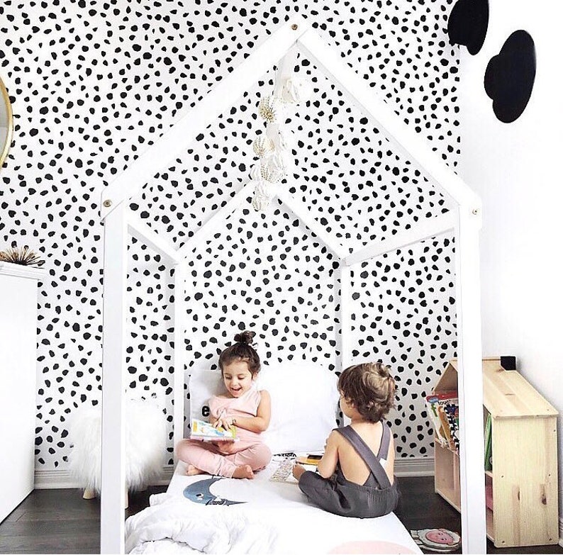 K&ouml;nnte beinhalten: Ein wei&szlig;er, hausf&ouml;rmiger Bettrahmen aus Holz mit zwei Kindern darin. Das Bett hat eine wei&szlig;e Bettdecke mit einem Mondsichel-Design. Der Hintergrund zeigt eine Wand mit einem schwarz-wei&szlig;en Dalmatiner-Muster. Ein kleines, hausf&ouml;rmiges B&uuml;cherregal aus Holz befindet sich rechts.