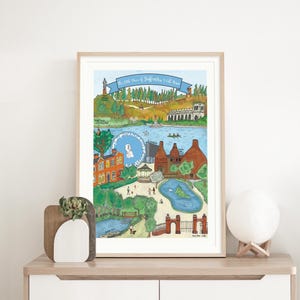 May include: A framed art print depicting a detailed, colourful illustration of a townscape with a lake, buildings, and trees. The print includes the text "The little place of Staffordshire I call home".