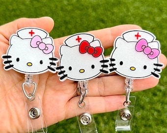 Pode incluir: Três porta-crachás de enfermeira Hello Kitty brancos com cruzes vermelhas e laços de diferentes cores. Os porta-crachás são presos a rolos retráteis de crachá transparentes.