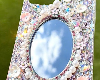 May include: A clear phone case with a mirror and rhinestones. The case is decorated with pink flowers, pearls, and iridescent crystals.