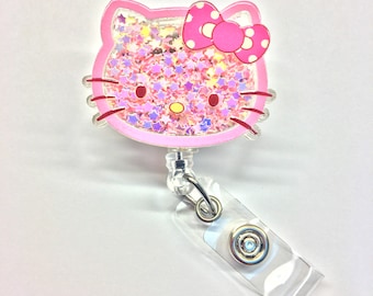 Pode incluir: Porta-crachá rosa com o personagem Hello Kitty com glitter e um laço rosa. O porta-crachá tem um carretel retrátil de plástico transparente.