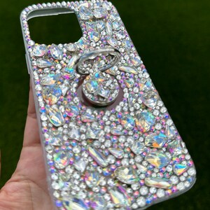 May include: A clear phone case covered in iridescent rhinestones with a silver ring holder attached.