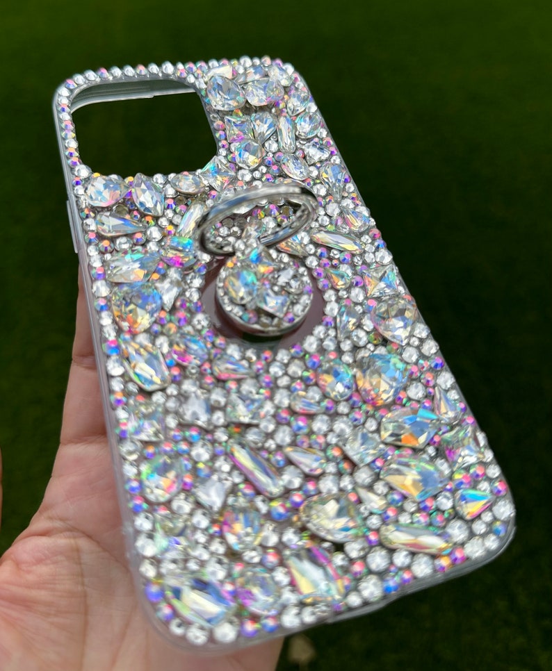 May include: A clear phone case covered in iridescent rhinestones with a silver ring holder attached.