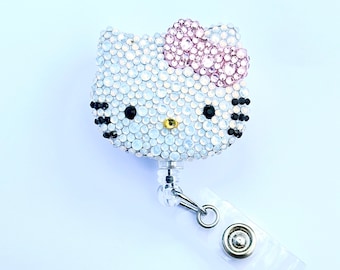 Pode incluir: Um porta-crachá retrátil Hello Kitty com strass. O rosto é coberto por strass transparentes, com um laço de strass rosa. Strass pretos formam os olhos e os bigodes, e um strass dourado é o nariz. Clipe de plástico transparente e fecho de metal estão anexados.