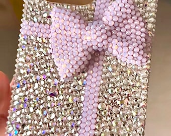 May include: A clear phone case covered in iridescent rhinestones with a large lavender bow in the center.