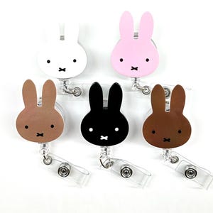 May include: A collection of six bunny-shaped retractable badge holders in various colors: white, pink, brown, black, and two shades of brown. Each badge holder features a simple bunny face design with a black 'x' mouth and two black dots for eyes.