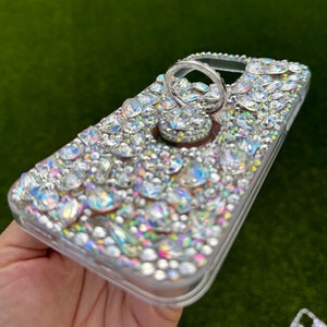 May include: A clear phone case covered in iridescent rhinestones with a silver ring holder attached.