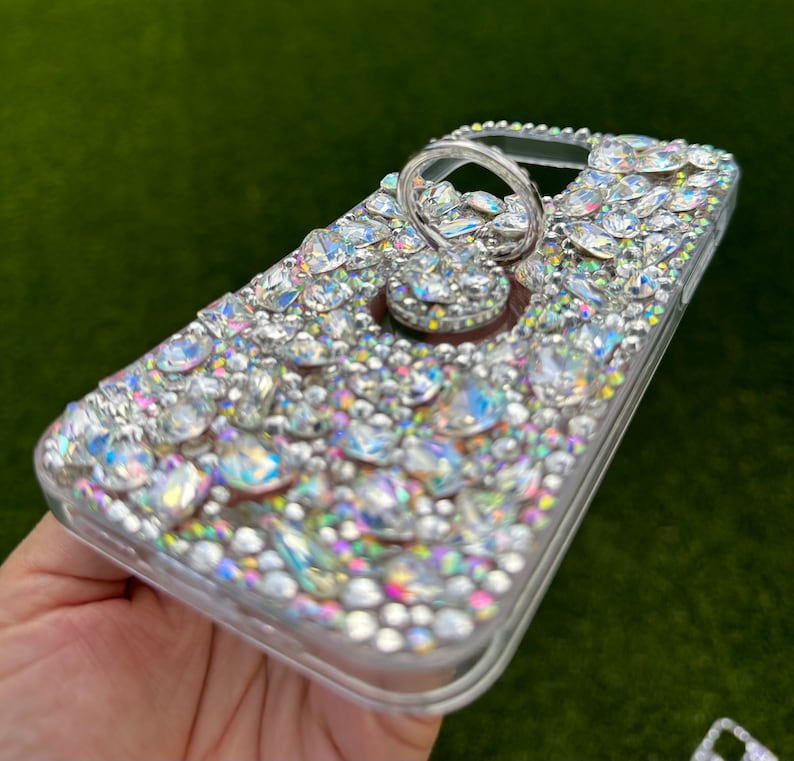 May include: A clear phone case covered in iridescent rhinestones with a silver ring holder attached.