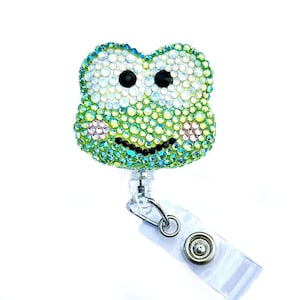 Puede incluir: Un portatarjetas con forma de rana adornado con pedrería. La rana está cubierta de pedrería iridiscente, con ojos negros y una boca sonriente. El portatarjetas tiene una correa de plástico transparente y un clip metálico.