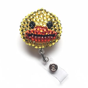 Può includere: Un portabadge con la faccia di un'anatra gialla, occhi neri e un becco rosso, fatto di strass. Il portabadge è attaccato a un porta badge in plastica trasparente.