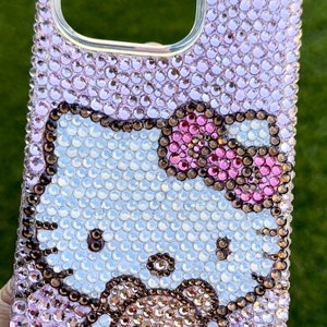 May include: A pink and white Hello Kitty phone case covered in rhinestones. The case features a Hello Kitty design with a pink bow and a brown bear.