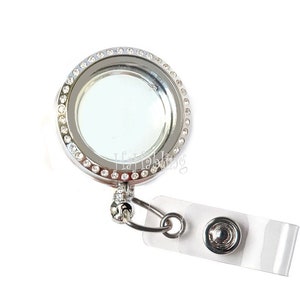 May include: Silver tone retractable badge holder with a clear round locket decorated with a row of clear rhinestones. The locket is attached to a clear plastic badge holder with a silver tone button.