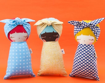 May include: Three handmade fabric dolls with different colored hair and clothing. The dolls are wearing headbands and are wrapped in blankets with different patterns. The dolls are on a bright orange background.