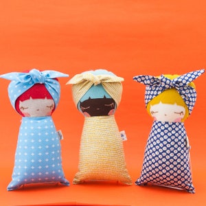 Puede incluir: Tres muñecas de tela hechas a mano con cabello y ropa de diferentes colores. Las muñecas llevan diademas y están envueltas en mantas con diferentes patrones. Las muñecas están sobre un fondo naranja brillante.