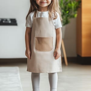 Può includere: Un grembiule beige con un'ampia tasca frontale, indossato sopra una maglietta bianca e leggings grigi. Il grembiule ha una tasca rettangolare con angoli arrotondati. Il testo "ithinksew" è nell'angolo in basso a destra. Il bambino indossa scarpe bianche.