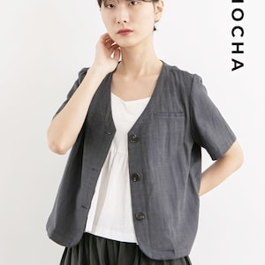 Puede incluir: Una camisa de lino gris de manga corta con cuello en V y un solo bolsillo en el lado izquierdo. La camisa está desabrochada y se ve una camiseta blanca debajo.