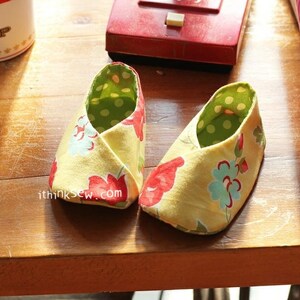 May include: A pair of yellow baby shoes with a floral print. The shoes have a green and white polka dot lining. The shoes are on a wooden surface.