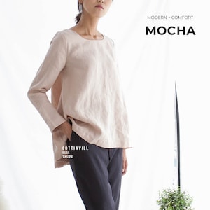 May include: A woman is wearing a light pink linen top with long sleeves and a scoop neck. She is also wearing dark gray cropped pants. The text "MODERN - COMFORT" and "MOCHA" is visible at the top of the image.