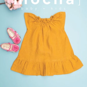 May include: Mustard yellow baby dress with a ruffled hem and a gathered neckline. The image also features pink floral shoes and a sprig of pink cotton. The text "mocha baby + kids" is displayed in white.