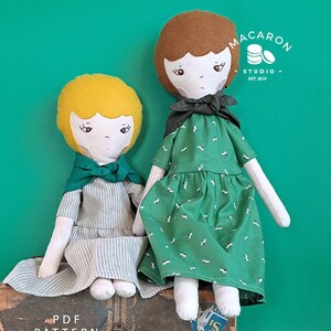 Puede incluir: Dos muñecas de tela hechas a mano con vestidos y pañuelos verdes. La muñeca de la izquierda tiene el pelo amarillo y un vestido a rayas. La muñeca de la derecha tiene el pelo castaño y un vestido verde con un estampado blanco. Las muñecas están sentadas en una maleta vintage. La imagen incluye el texto "Macaron Studio - Est. 2018" y "PDF Pattern Modern Design".