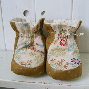 Könnte beinhalten: Ein Paar braune Babyschuhe mit weißem Blumenmuster. Die Schuhe haben einen gerafften elastischen Bund und sind aus weichem Stoff.