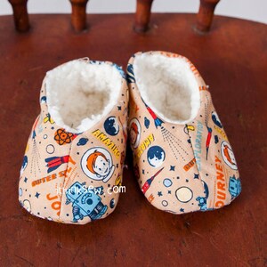 May include: A pair of beige slipper booties with a space-themed print featuring rockets, planets, stars, and astronauts. The booties have a white fleece lining.