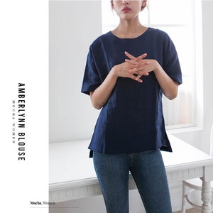 May include: A navy blue linen blouse with a relaxed fit and a slightly longer hem at the back. The blouse has short sleeves and a round neckline. The model is wearing the blouse with blue jeans.