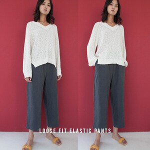 May include: Two images of a person wearing a cream-colored, loose-fitting V-neck sweater and gray, wide-leg pants. The text "LOOSE FIT ELASTIC PANTS" is at the bottom of the image. The background is a solid red.