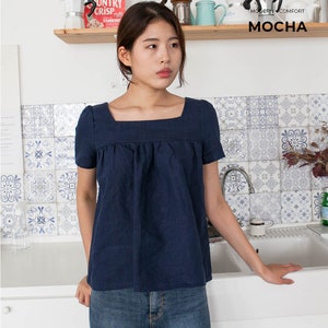 May include: A woman wearing a navy blue square neck top with short sleeves and blue jeans. The top has a gathered detail at the bust. The woman is standing in a kitchen with white cabinets and a tiled backsplash.