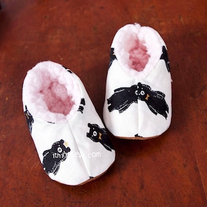 May include: A pair of white baby booties with a black bear print and pink fleece lining. The booties are sitting on a wooden chair.