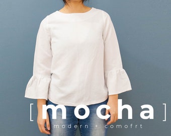May include: A white long-sleeve blouse with bell sleeves and a round neckline. The blouse is worn over blue jeans. The text "[mocha] modern + comfort" is printed below the image.