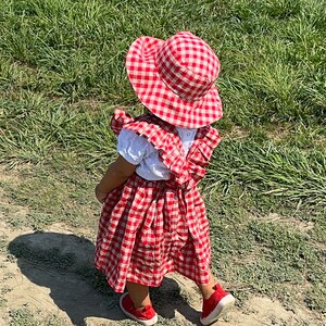 May include: A young child wearing a red and white gingham dress and matching sun hat. The dress has a white ruffle trim and the child is wearing red shoes.