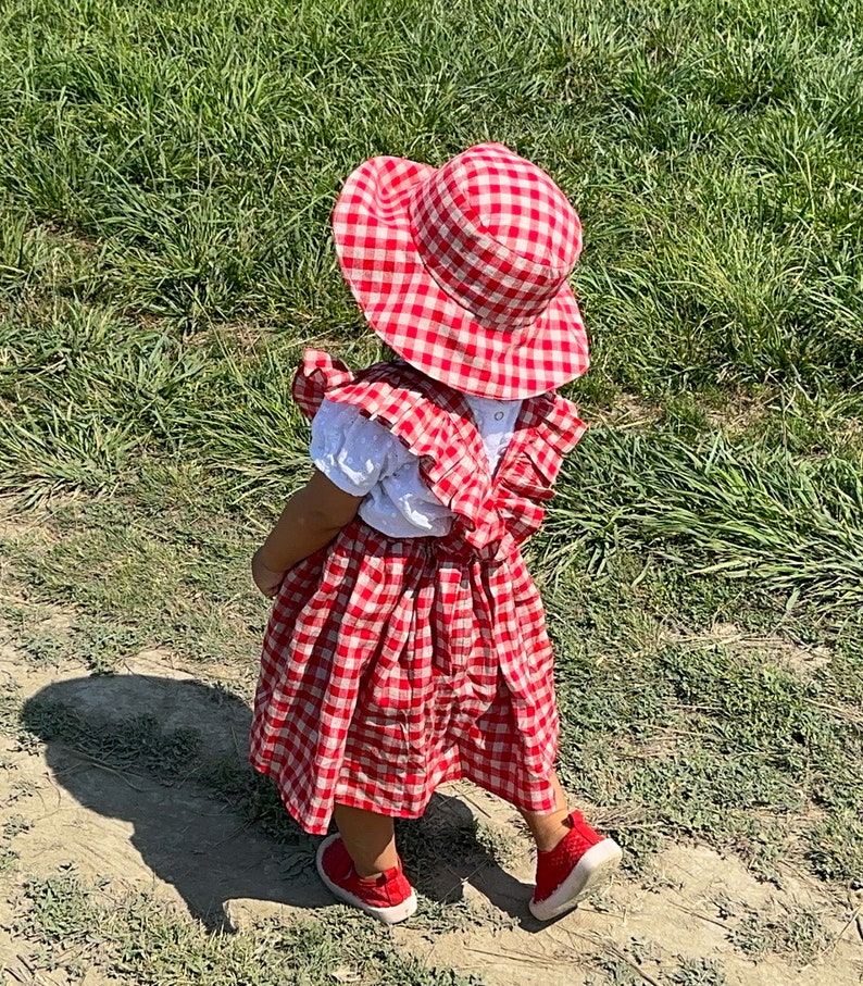 May include: A young child wearing a red and white gingham dress and matching sun hat. The dress has a white ruffle trim and the child is wearing red shoes.