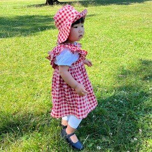 May include: A young child wearing a red and white gingham dress and a red and white gingham sun hat stands on a grassy lawn. The child is wearing black shoes and white tights.