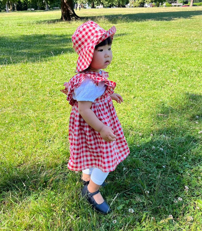 May include: A young child wearing a red and white gingham dress and a red and white gingham sun hat stands on a grassy lawn. The child is wearing black shoes and white tights.