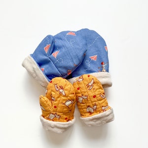 May include: A pair of yellow baby mittens with a white trim and a pattern of small white animals. The mittens are paired with a blue hat with a white trim and a pattern of red paper aeroplanes.