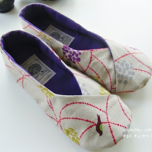 May include: A pair of beige fabric slippers with a floral pattern in red, yellow, and purple. The interior is a deep purple, and each slipper has a label. The slippers are designed for comfort and indoor use.