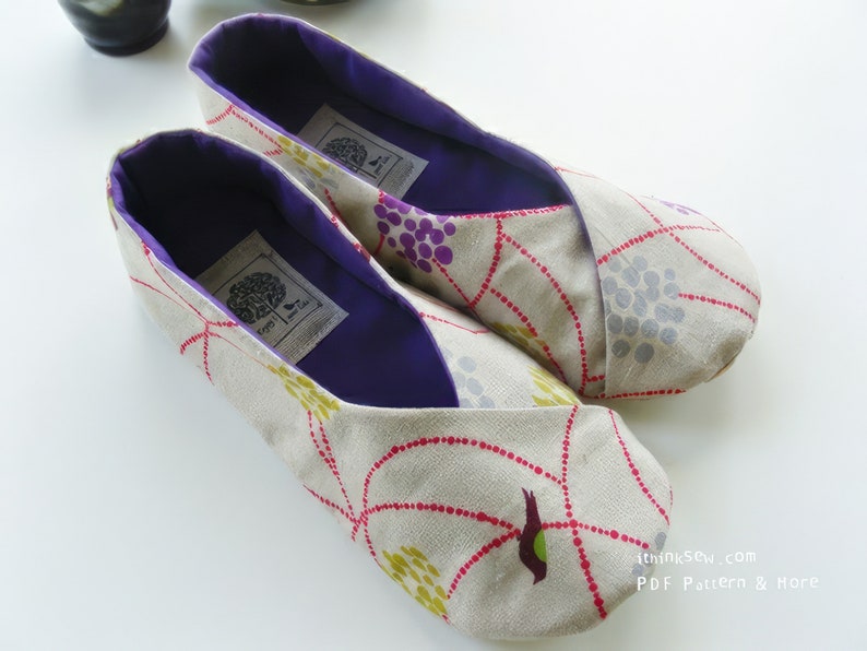 May include: A pair of beige fabric slippers with a floral pattern in red, yellow, and purple. The interior is a deep purple, and each slipper has a label. The slippers are designed for comfort and indoor use.
