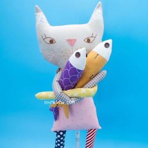 May include: A white cat plush toy with embroidered eyes and a pink nose, holding two fish plush toys. One fish is purple with white polka dots and the other is yellow with brown spots. The cat is wearing a pink dress with white polka dots and red and white striped legs. The text "ithinksew.com" is printed on the bottom of the image.