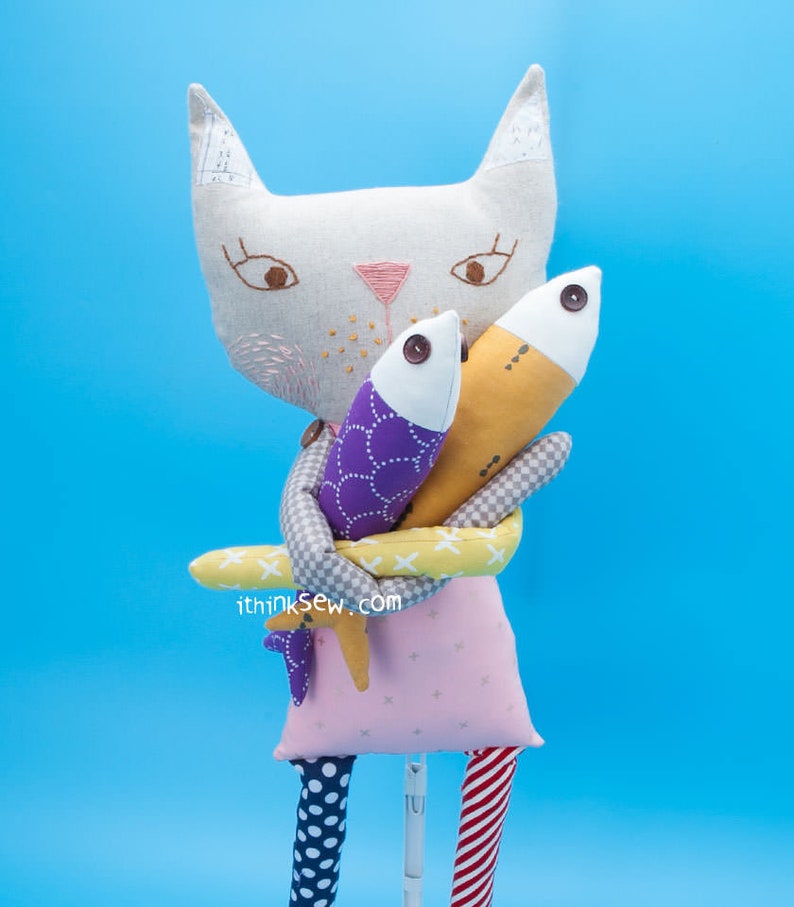 May include: A white cat plush toy with embroidered eyes and a pink nose, holding two fish plush toys. One fish is purple with white polka dots and the other is yellow with brown spots. The cat is wearing a pink dress with white polka dots and red and white striped legs. The text "ithinksew.com" is printed on the bottom of the image.