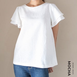 May include: White linen top with a round neckline and double ruffle short sleeves. The top is loose fitting and falls to the hips. The model is wearing blue jeans.