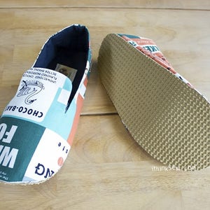 May include: A pair of blue, green, and white fabric slippers with a brown sole. The slippers have a graphic print of a chocolate bar with the text "Choco-Band" and "Walking for..." on the front.
