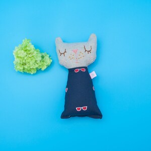 May include: A gray cat plush toy with embroidered eyes and a red heart on its chest. The cat is wearing a blue dress with red heart accents and a pink tag that says "The Little" on it.