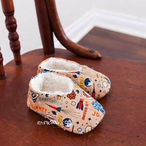 May include: A pair of beige baby slippers with a space-themed print. The print features rockets, planets, stars, and the words "Journey", "Exploration", "Outer Space", and "Atomic".