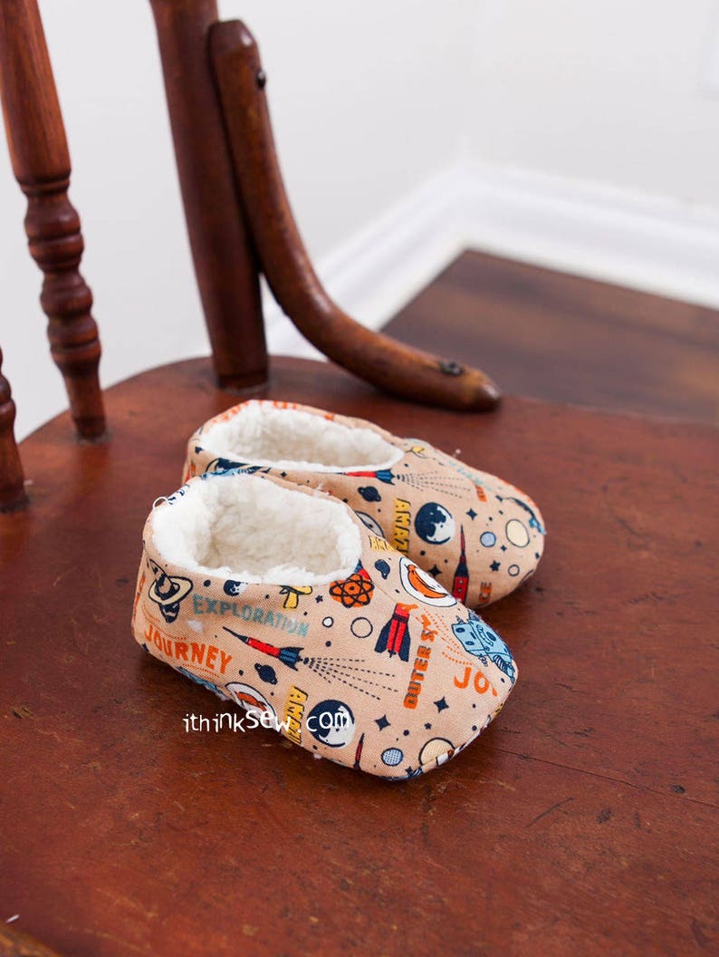 May include: A pair of beige baby slippers with a space-themed print. The print features rockets, planets, stars, and the words "Journey", "Exploration", "Outer Space", and "Atomic".