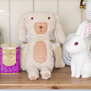 May include: A white plush toy dog with brown eyes and a stitched brown nose. The dog is sitting on a wooden surface with a box of tea and a white plush toy rabbit beside it.
