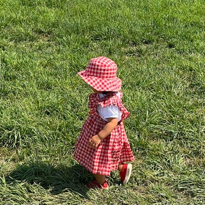 May include: A young child wearing a red and white gingham dress and a red and white gingham hat walks on a grassy field.