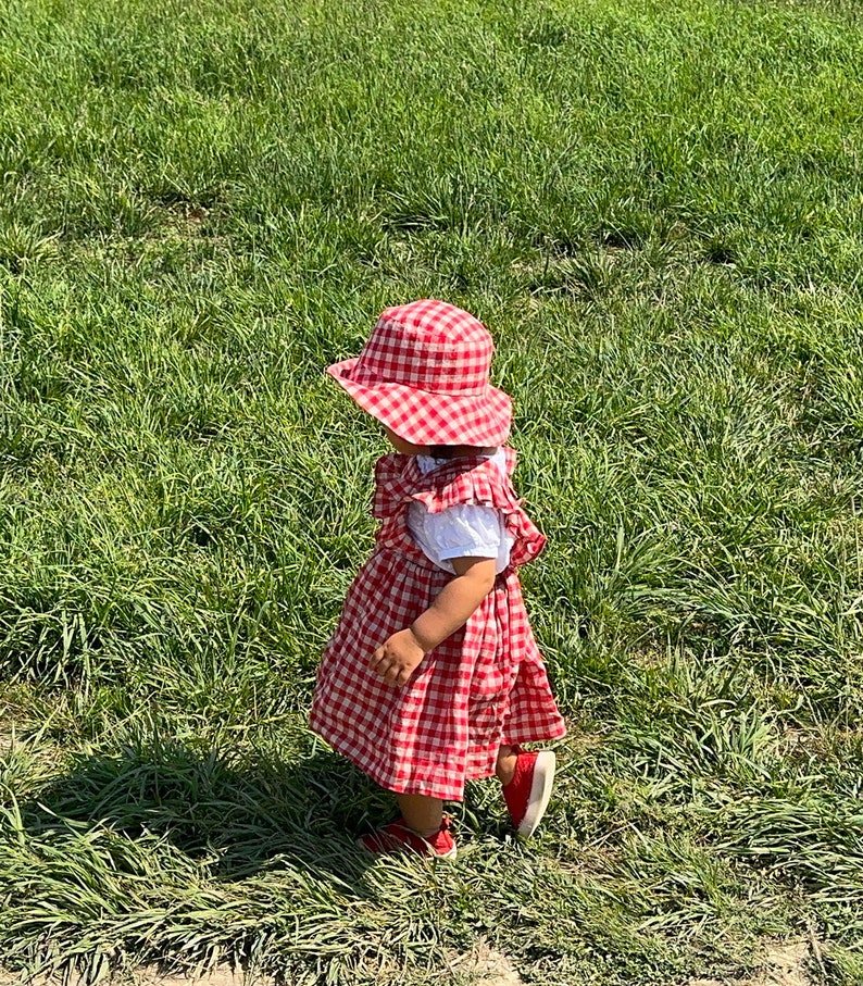 May include: A young child wearing a red and white gingham dress and a red and white gingham hat walks on a grassy field.